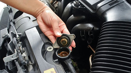 Hand holding a car radiator cap above the engine, illustrating vehicle cooling system maintenance and repair.