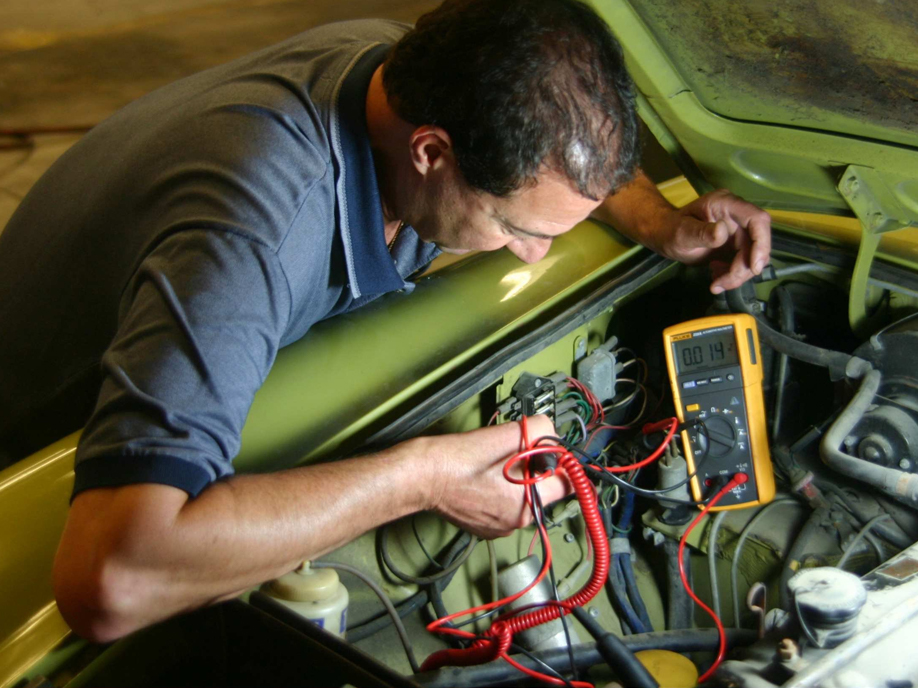 Technician diagnosing vehicle electrical system with multimeter, inspecting wiring and connections under the hood, emphasizing electrical repair services in Columbus, OH.