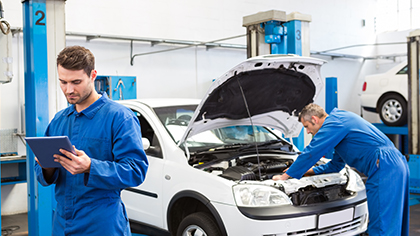 Mechanics in blue coveralls working on a vehicle in an auto repair shop, one using a tablet for diagnostics while the other inspects the engine, representing preventive maintenance services.